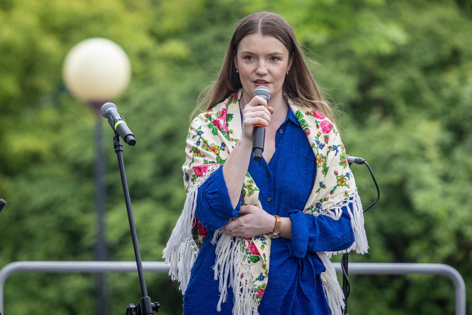 Zdjęcie. Kobieta o długich prostych włosach mówi do mikrofonu. Na sobie ma niebieską sukienkę i białą chustę w kwiaty zarzuconą na ramiona. To dyrektor KCK Agata Klimczak-Kołakowska.