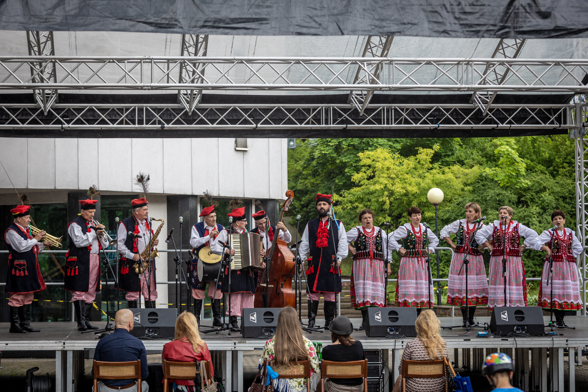 Zdjęcie. Plenerowa scena KCK. Na niej zespół ludowy. Kobiety w błyszczących gorsetach, kwiecistych spódnicach i przezroczystych fartuchach trzymają się pod boki. Po lewej stronie stoi zespól muzyczny. Panowie mają na głowie czerwone rogatywki, a w dłoniach instrumenty. Na środku stoi mężczyzna z brodą w długiej kamizelce sięgającej aż do kolan, tzw. sukmanie z czerwonymi pędzelkami przy zapięciu.