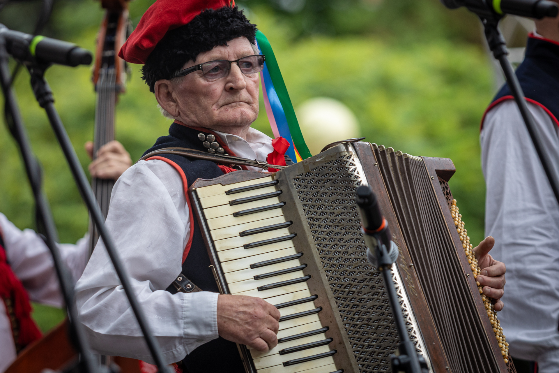 Zdjęcie. Starszy mężczyzna w okularach i czerwonej rogatywce gra na starej harmonii w brązowym kolorze z pożółkłymi ze starości klawiszami.