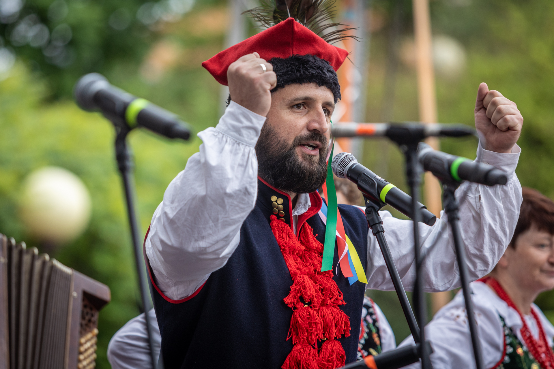 Zdjęcie. Mężczyzna w białej koszuli, granatowej kamizelce z czerwonymi pędzelkami i czerwonej czapce rogatywce z pawim piórem stoi przed mikrofonem. Obie ręce unosi do góry i ściska w pięść. Mężczyzna ma ciemne wąsy i brodę.