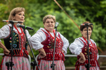 Zdjęcie. Trzy kobiety w haftowanych w kwiaty gorsetach, białych bluzkach i czerwonych koralach na szyi stoją w rzędzie. Wszystkie trzymają się pod boki.
