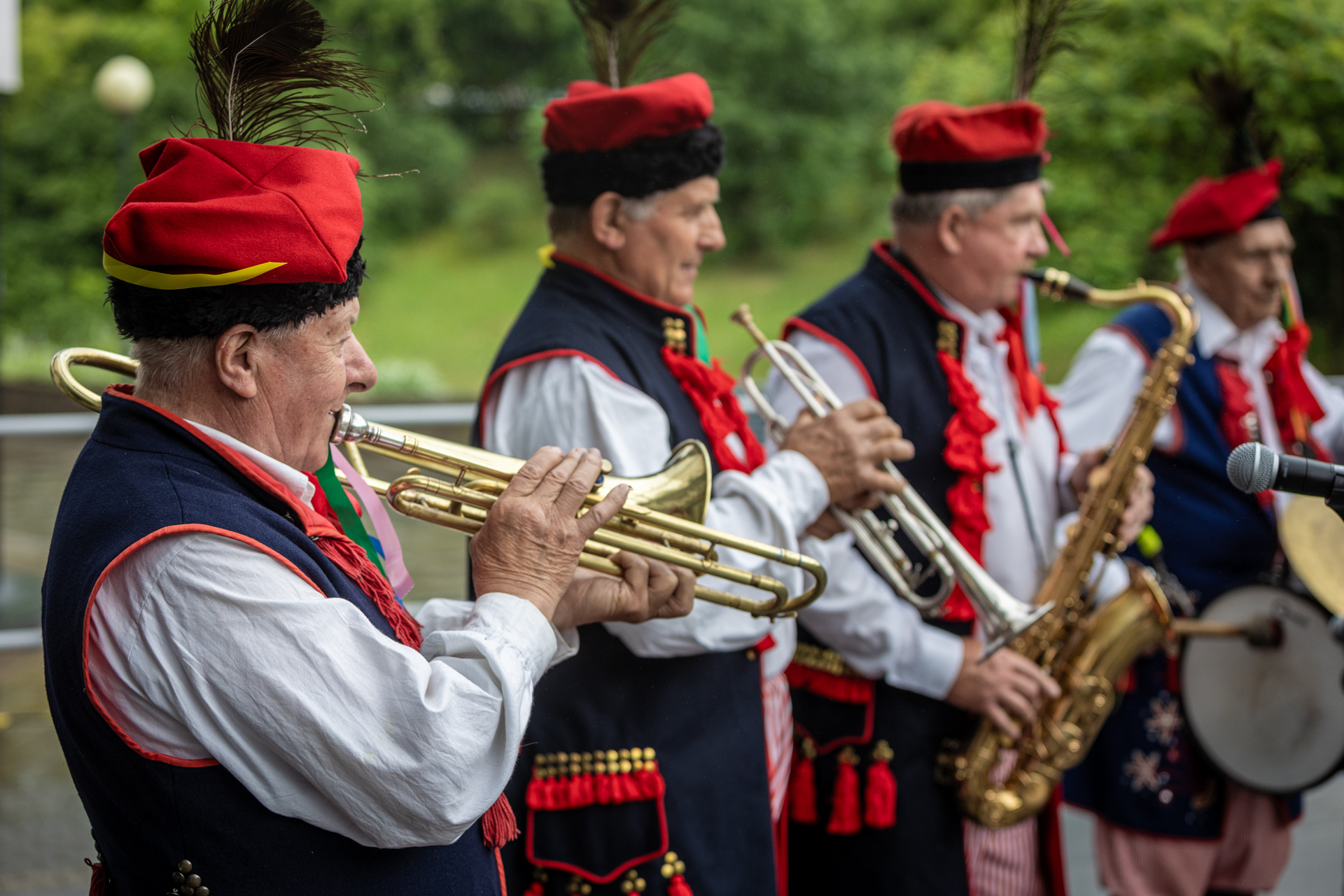 Zdjęcie. Czterech mężczyzn w granatowych kamizelkach i czerwonych rogatywkach na głowie gra na instrumentach.