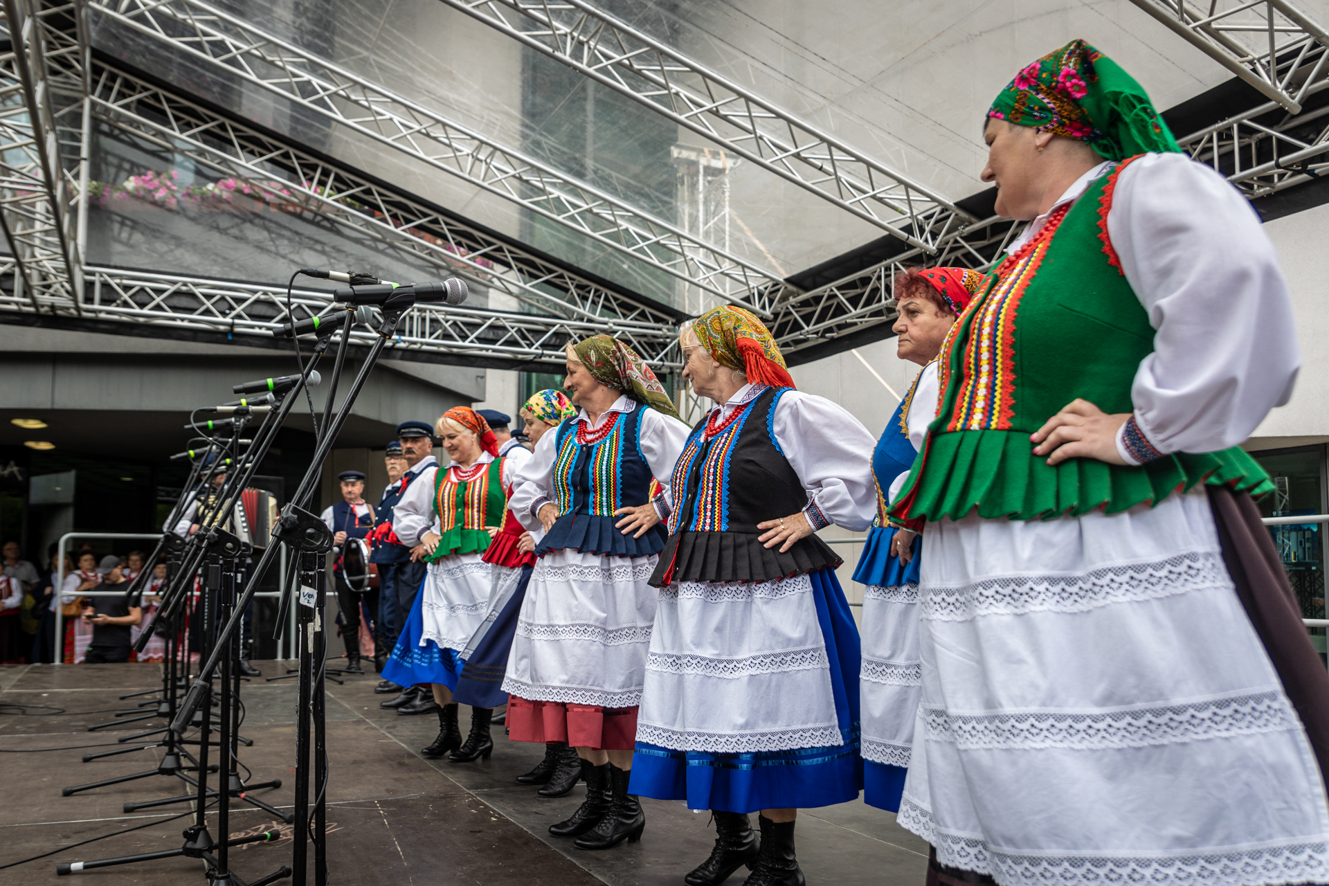 Zdjęcie. Kobiety w chustkach na głowie, kolorowych gorsetach z paskami pionowymi przy zapięciach, jednokolorowymi spódnicami i białymi fartuchami z koronką stoją w rzędzie na scenie. Wszystkie trzymają się pod boki.