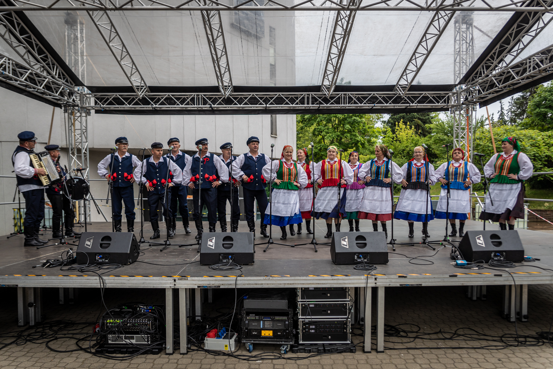 Zdjęcie. Zespół ludowy na scenie letniej KCK. Po prawej stronie w rzędzie stoją kobiety w kolorowych gorsetach, jednokolorowych spódnicach i chustkach na głowie. Po lewej stronie stoją panowie w niebieskich spodniach, kamizelkach i czapkach z daszkiem na głowie. Na krawędzi sceny stoi dwóch muzyków. Akordeonista i perkusista.