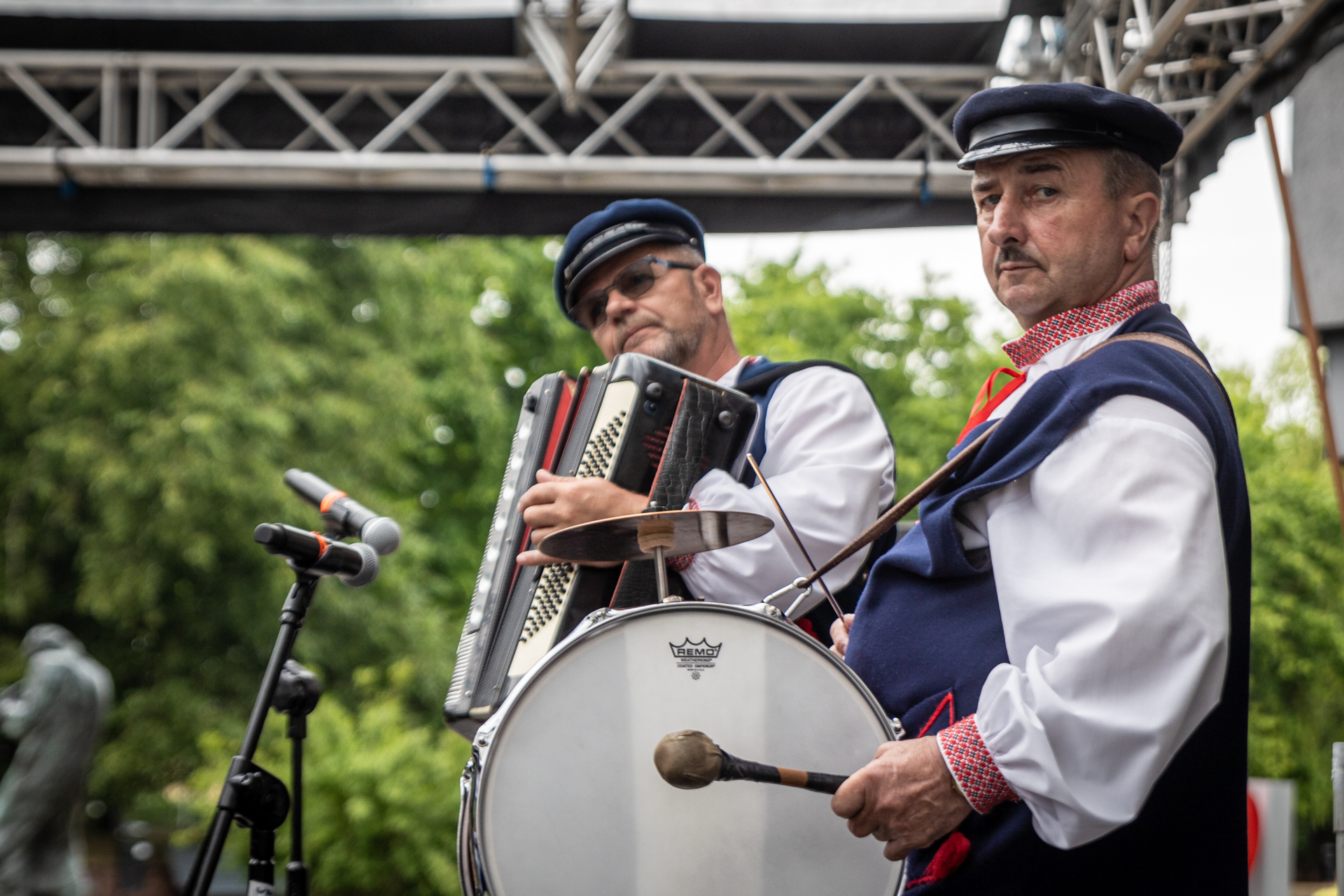 Zdjęcie. Dwaj mężczyźni w granatowych czapach z daszkami grają na instrumentach> Jeden na harmonii, drugi na bębnie i talerzy perkusyjnym zawieszonym na szyi.