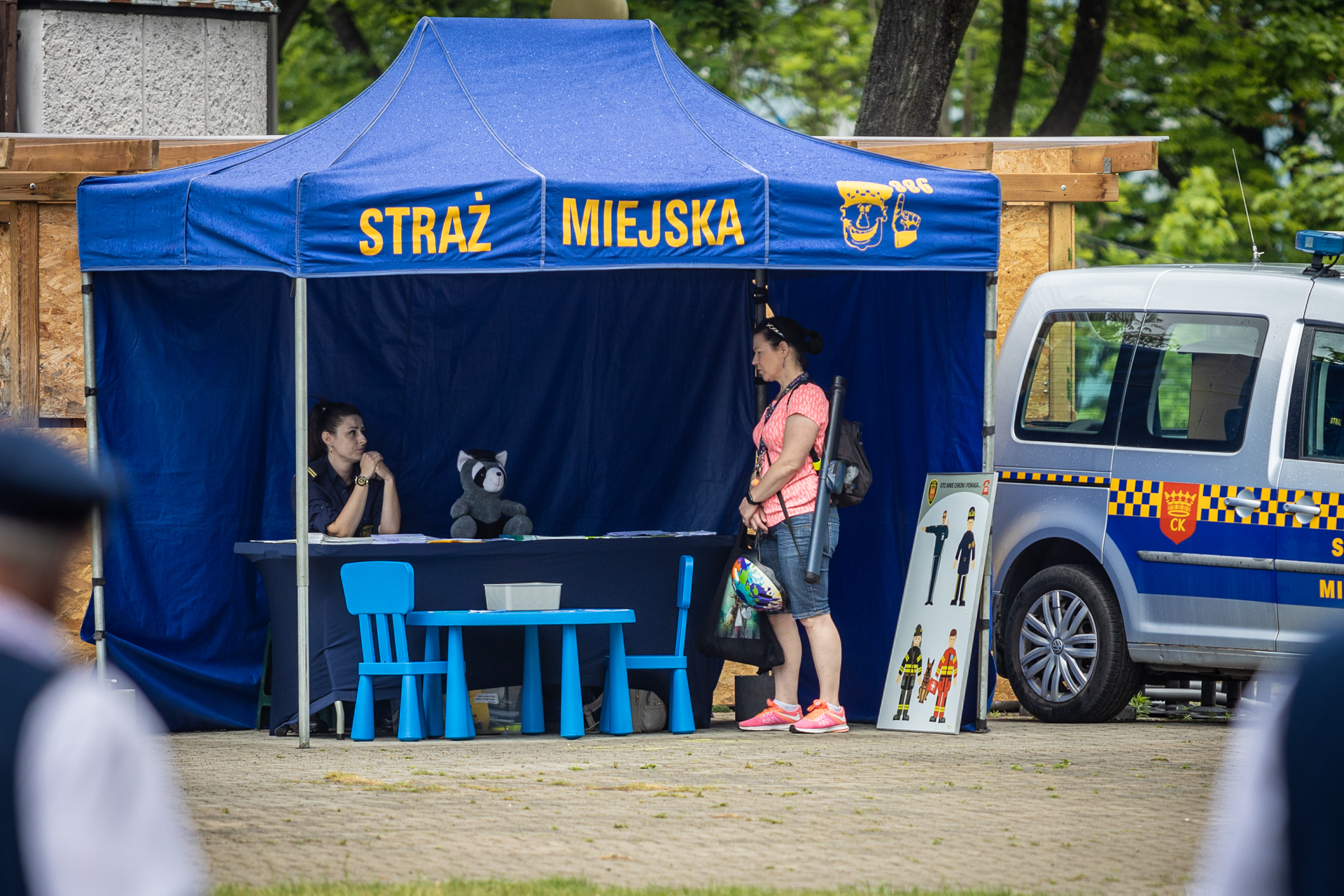 Zdjęcie. Niebieski namiot ze stołem. Za stołem siedzi kobieta w mundurze. Na górze namiotu żółty duży napis: Straż Miejska.