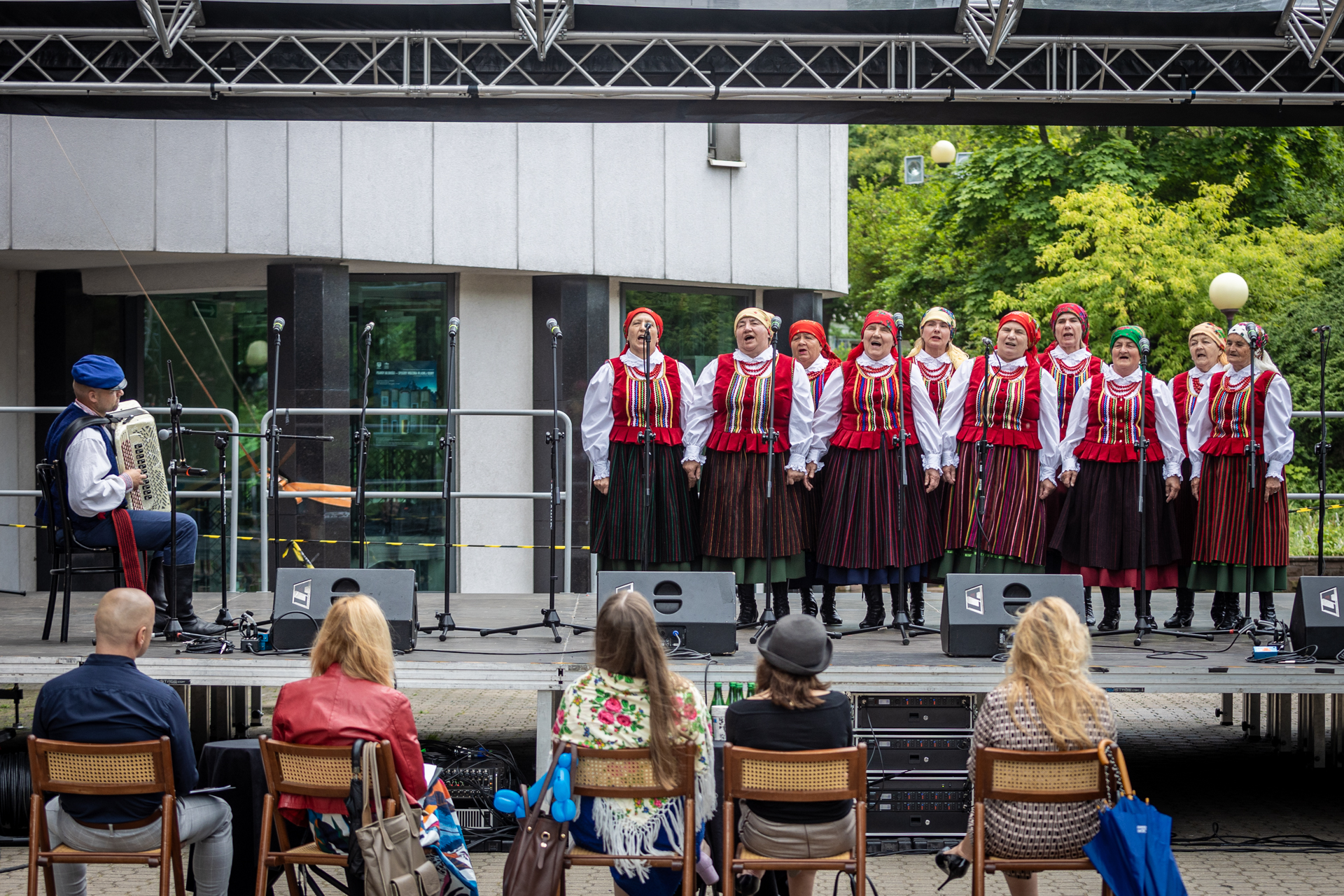 Zdjęcie. Dziesięć kobiet w strojach ludowych stoi na scenie i śpiewa do mikrofonu. Obok mężczyzna akompaniuje im na harmonii. Kobiety mają na sobie czerwone gorsety z ozdobami w kolorowe, pionowe paski i czarne spódnice w wąskie, kolorowe paski. Na głowach mają kolorowe chusty w kwiaty.