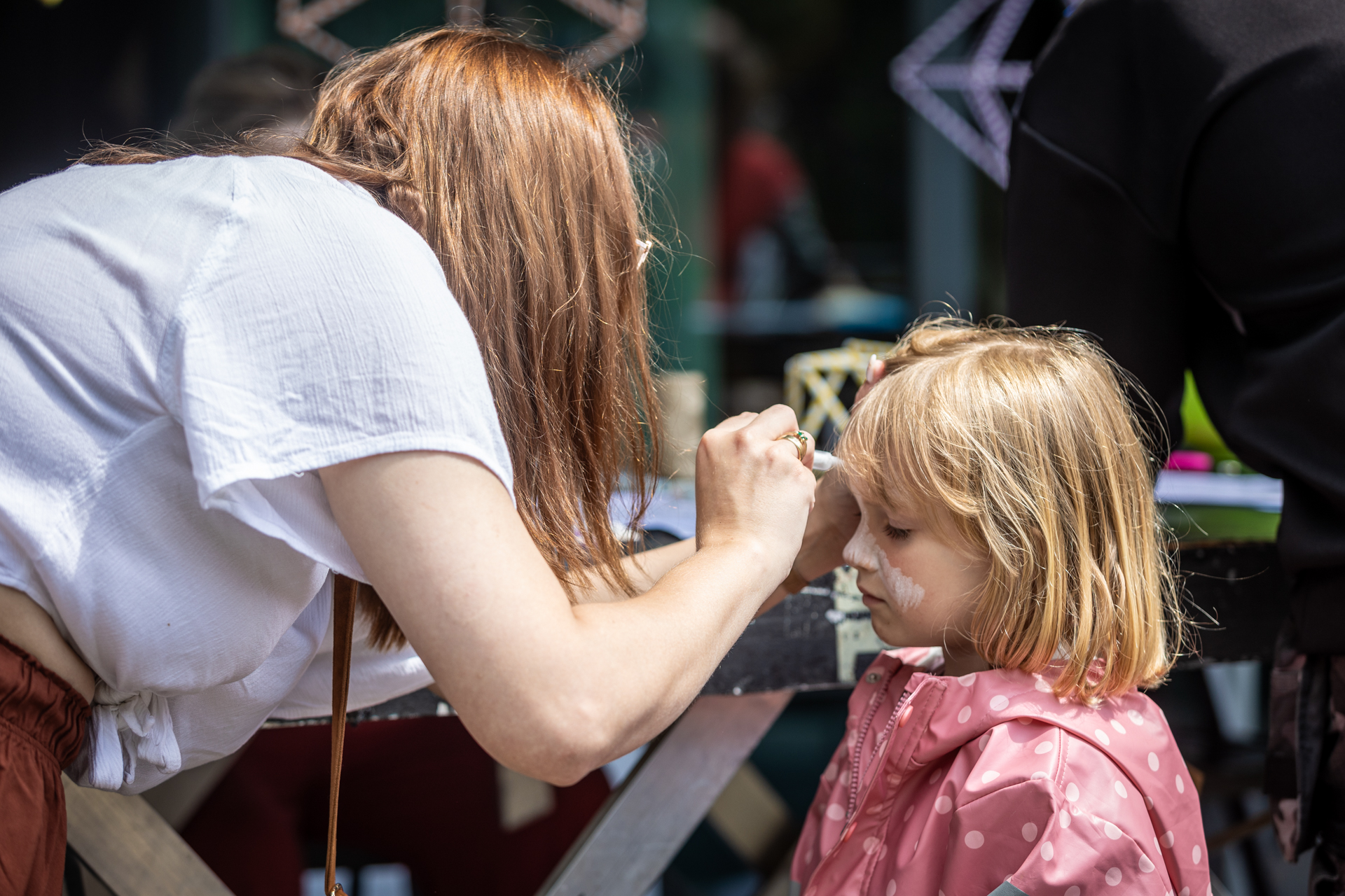 Zdjęcie. Kobieta o jasnych włosach maluje kredką buzię niewielkiej dziewczynki o blond włosach do ramion. Dziewczynka ma na sobie kurtkę przeciwdeszczową w kolorze różowym z dużymi białymi kropkami.