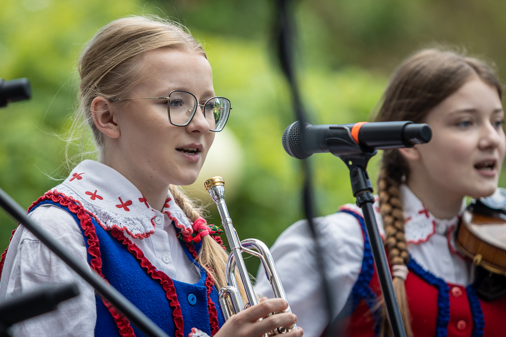 Zdjęcie. Dwie dziewczynki śpiewają. Blondynka ma na nosie okulary i włosy zaplecione w warkocz. W ręku trzyma trąbkę. Druga ma dwa ciemniejsze warkocze i skrzypce.