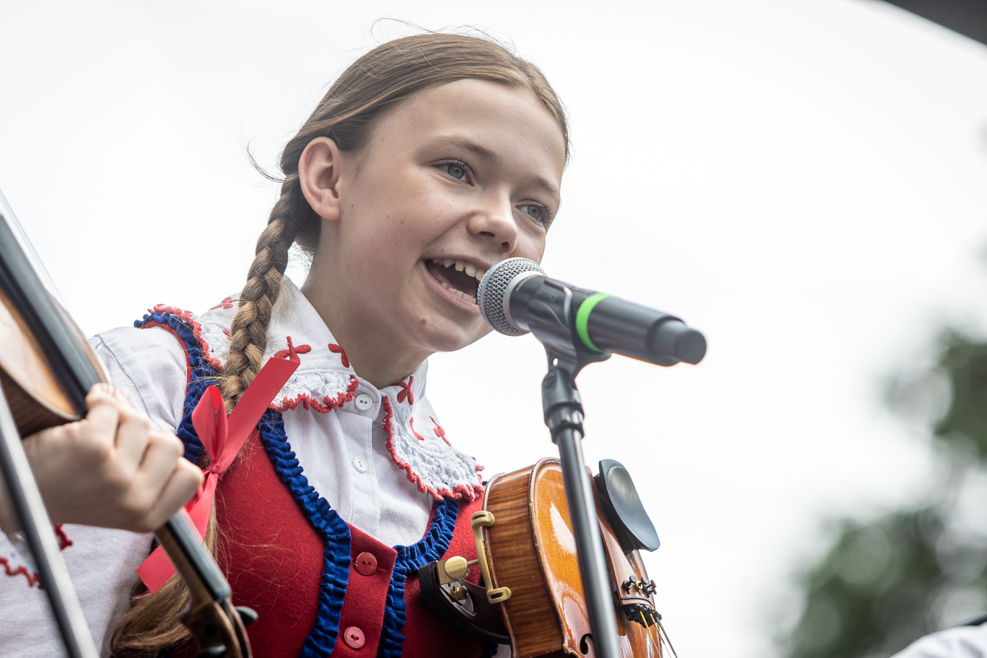 Zdjęcie. Dziewczynka z dwoma warkoczami ubrana w białą bluzkę z dużym zakończony na czerwono kołnierzem i czerwony gorset z niebieskimi drobnymi falbankami śpiewa do mikrofonu.