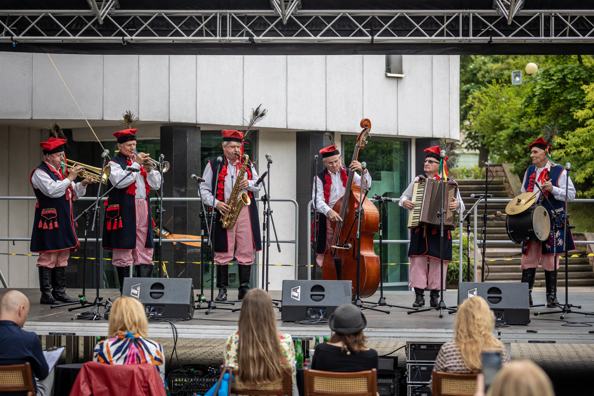 Zdjęcie. Sześciu mężczyzn w krakowskich strojach ludowych na scenie. Wszysy grają na instrumentach: trąbkach, saksogfonie, harmonii, kontrabasie i perkusji zawieszonej przez jednego muzyka na pasku na szyi.