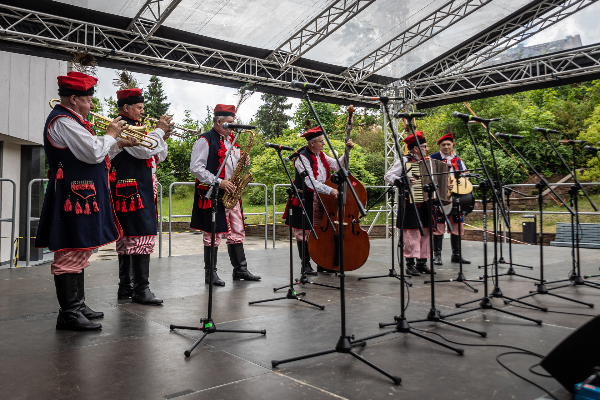 Zdjęcie. Sześciu mężczyzn w krakowskich strojach ludowych na scenie. Wszyscy grają na instrumentach: trąbkach, saksofonie, harmonii, kontrabasie i perkusji zawieszonej przez jednego muzyka na pasku na szyi.