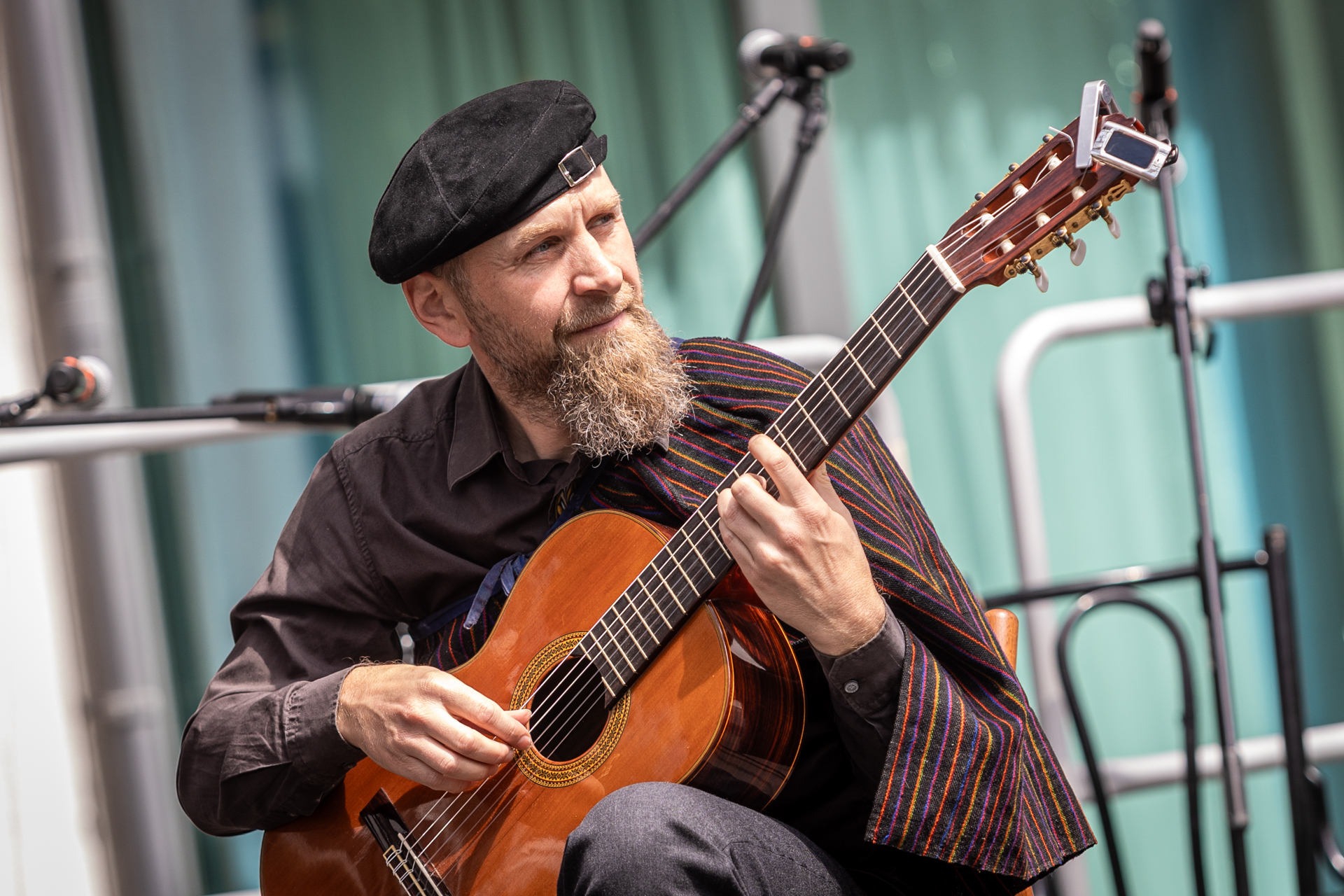 Zdjęcie. Mężczyzna z długa brodą z zapaską zarzuconą na ramię gra na gitarze. Na głowie ma czarną czapkę lub beret założony tyłem do przodu. Nad czołem ma sprzączkę pozwalającą dopasować nakrycie głowy do jej rozmiarów.