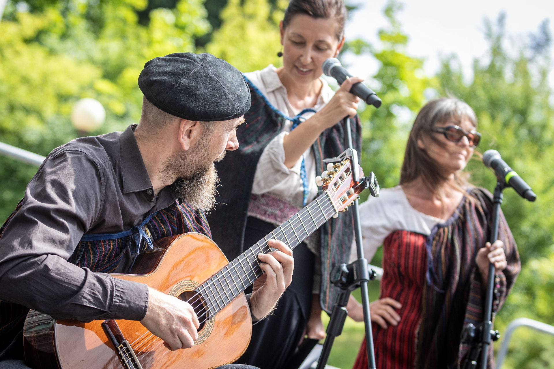 Zdjęcie. Trójka ludzi występuje na scenie. To dwie kobiety stojące przed mikrofonami i mężczyzna w czarnym berecie, który gra na gitarze.
