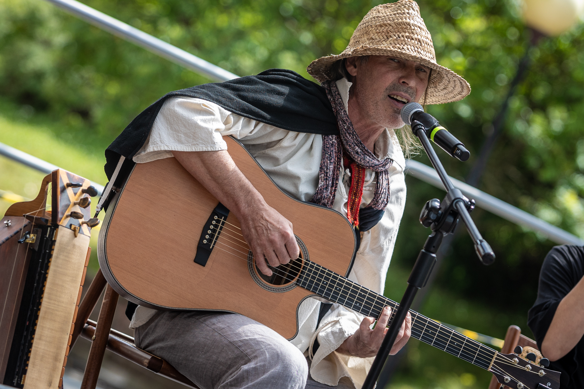 Zdjęcie. Mężczyzna w zapasce i słomkowym kapeluszu śpiewa do mikrofonu, jednocześnie grając na gitarze.