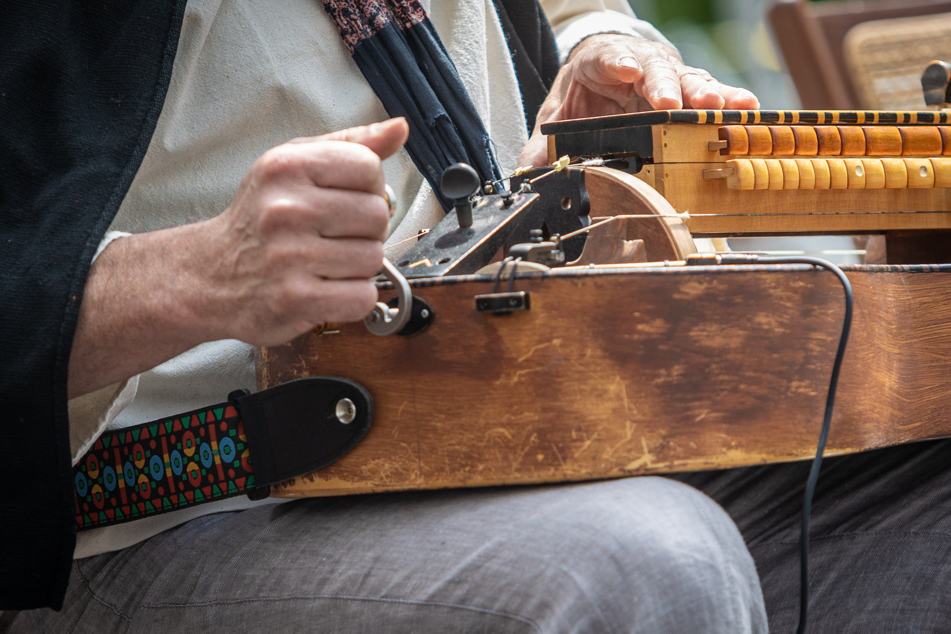Zdjęcie. Dłonie siedzącego muzyka. Na kolanach mężczyzna trzyma instrument. Widoczne jest duże drewniane pudło, a na nim drewniana klawiatura. Pudło zakończone jest metalowym hakiem. Mężczyzna trzyma lewą ręką ten hak. Prawą ręką gra na klawiaturze.
