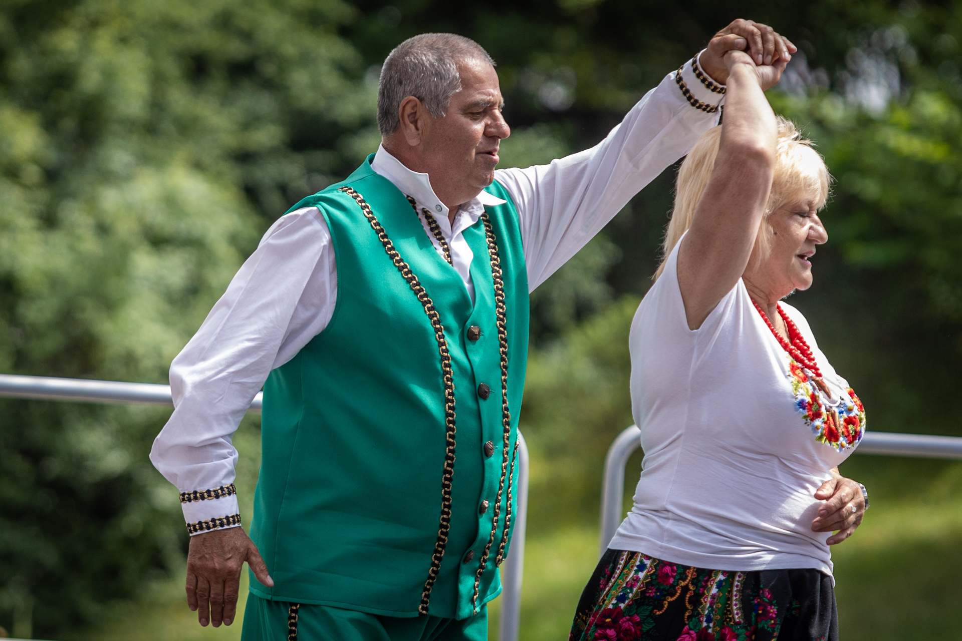 Zdjęcie. Mężczyzna w seledynowej kamizelce nałożonej na białą koszulę tańczy z kobietą w białej podkoszulce z kolorowym haftem we wzór kwiatów. Mężczyzna podnosi lewą rękę do góry, kobieta trzyma go prawą ręką i wykonuje obrót dookoła własnej osi.