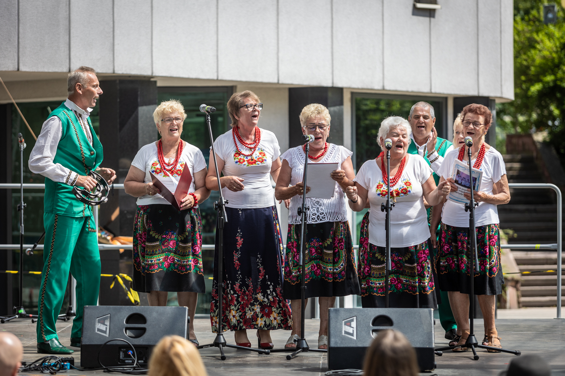 Zdjęcie. Sześć kobiet i dwóch mężczyzn śpiewa na scenie. Kobiety mają na sobie białe bluzki z kwiatowymi haftami i czerwone korale na szyi. Mają także czarne spódnice w kwiatowe ludowe wzory. Mężczyźni ubrani są w zielone spodnie i kamizelki.