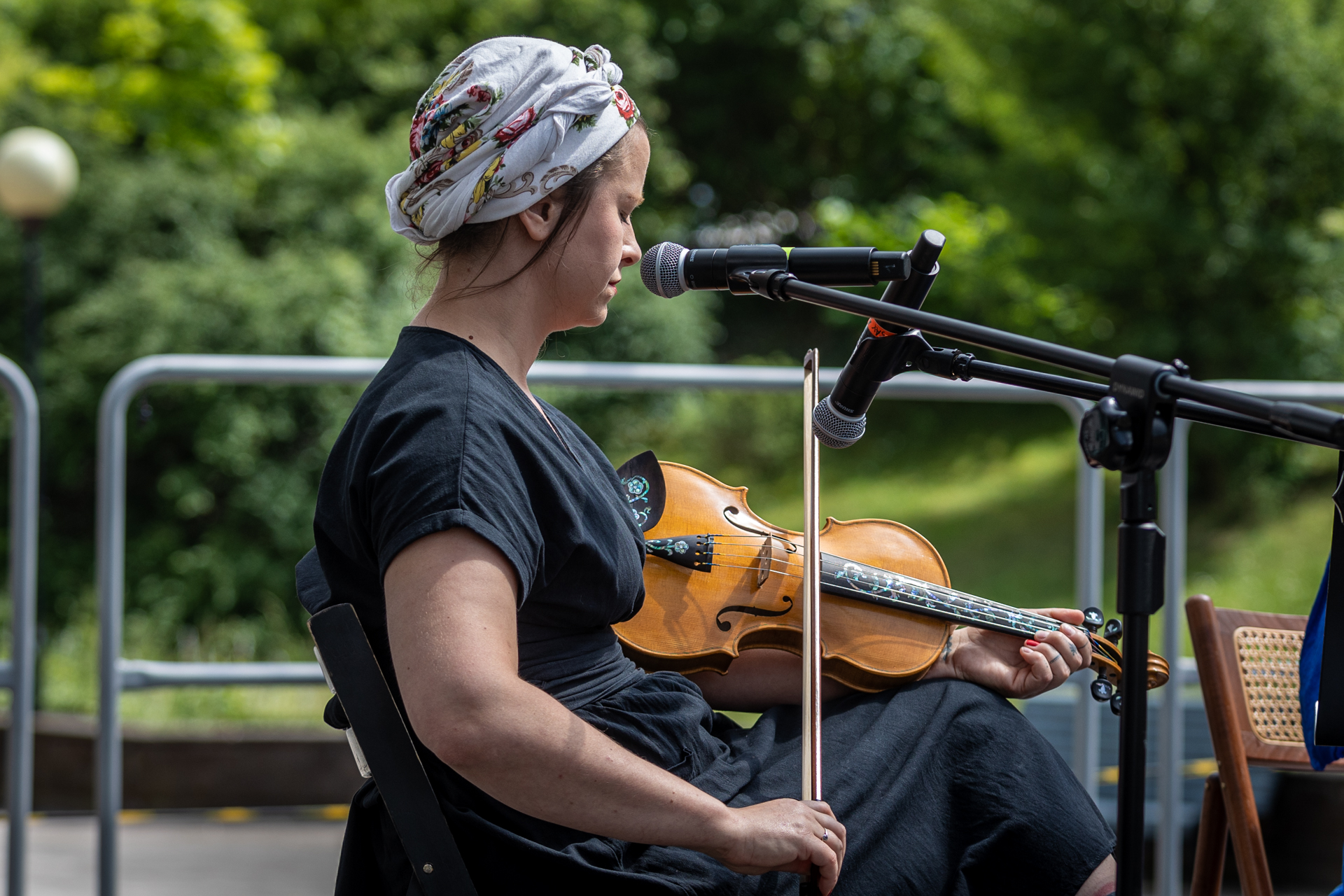 Zdjęcie. Kobieta w ciemnej sukience i jasnej kwiecistej chuście na głowie zawiniętej w turban gra na skrzypcach. Trzyma j nie pod brodą, ale na wysokości pachy.
