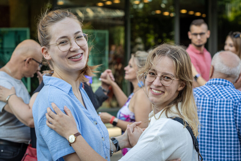 Zdjęcie. Dwie blondynki w okularach tańczą w parze. Obie odwracają się w kierunku aparatu i uśmiechają się.
