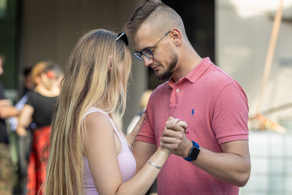 Zdjęcie. Młodzi ludzie tańczą w parze. Mężczyzna ma na sobie koszulkę polo w różowym kolorze. Kobieta o długich blond włosach jasnoróżową podkoszulkę na ramiączkach. Oboje patrzą w dół.
