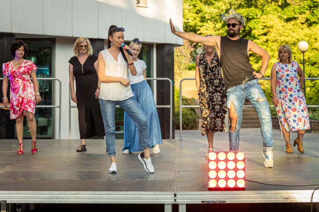 Zdjęcie. Grupa ludzi stoi na scenie. Przed nimi kobieta z mikrofonem. Wszyscy uginają lewą nogę w kolanie. W pierwszym rzędzie mężczyzna wyciąga prawą wyprostowaną rękę w bok.