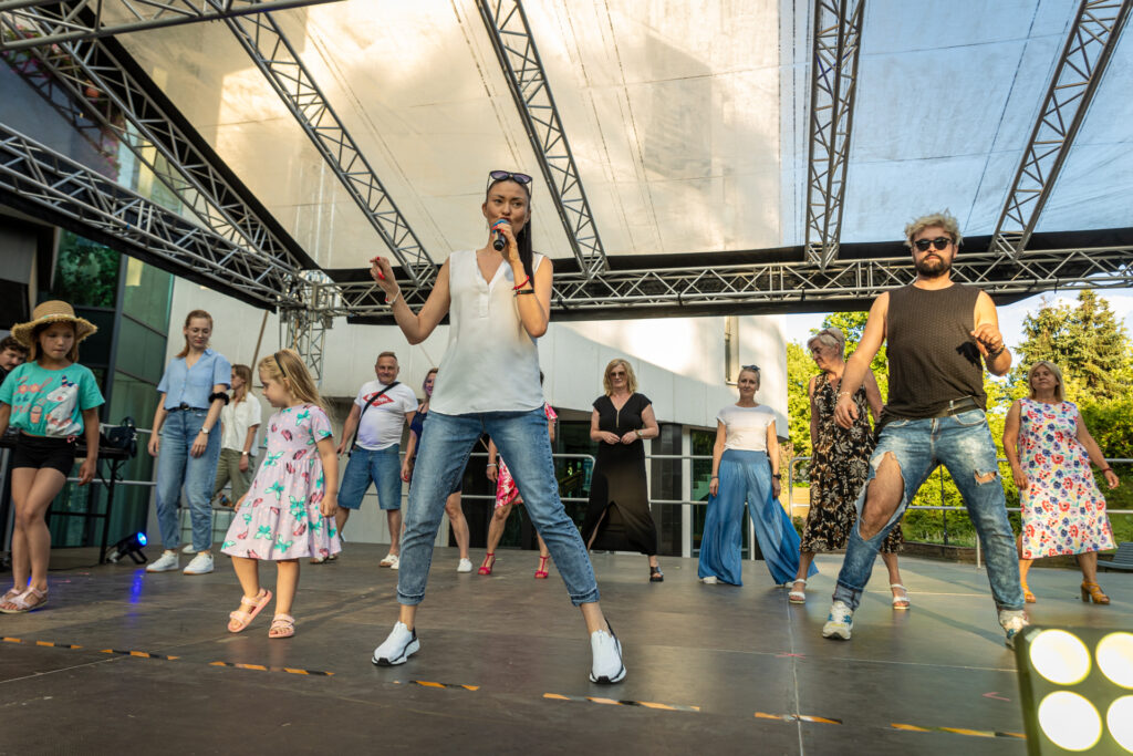 Zdjęcie. Grupa ludzi na scenie. W pierwszym rzędzie brunetka z mikrofonem. To Kamila Drezno. Wszyscy stoją w rozkroku.