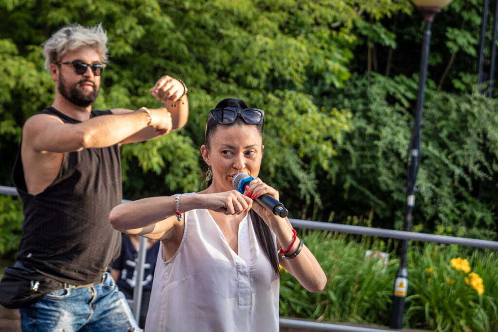 Zdjęcie. Dwójka ludzi. Drobna brunetka trzyma w lewej ręce mikrofon, prawą zgiętą w łokciu wyciąga przed siebie. Za nią mężczyzna z jasną grzywką naśladuje ruch rąk kobiety.