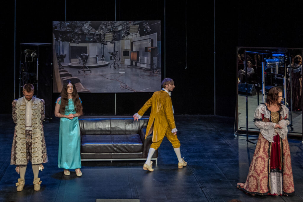 Zdjęcie. Mała scena KCK, na której stoją cztery osoby. W tle ekran ze zdjęciem studia telewizyjnego. Aktorzy teatru Pegaz są ubrani w stroje historyczne. Mężczyźni w żupany, spodnie do kolan z pończochami. Kobiety w długie suknie. Jeden z mężczyzn wychodzi ze sceny.