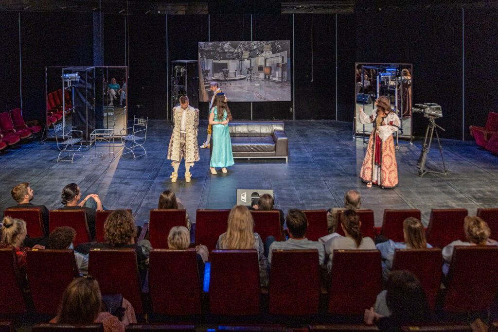 Zdjęcie. Mała scena KCK, na której stoją cztery osoby. W tle ekran ze zdjęciem studia telewizyjnego. Aktorzy teatru Pegaz są ubrani w stroje historyczne. Mężczyźni w żupany, spodnie do kolan z pończochami. Kobiety w długie suknie. Jeden z mężczyzn patrzy w podłogę. Przed aktorami widzowie spektaklu siedzą na czerwonych fotelach.