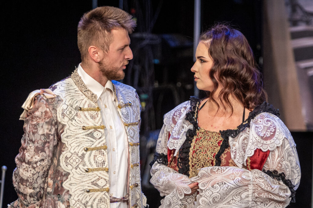 Zdjęcie. Dwójka młodych aktorów ubranych w historyczne stroje patrzy na siebie na scenie. Mężczyzna ma wzorzysty, wykończony koronkami surdut. Kobieta ma suknię z koronkowymi bufiastymi rękawami i bordowym, zdobionym złotą nitką gorsetem.