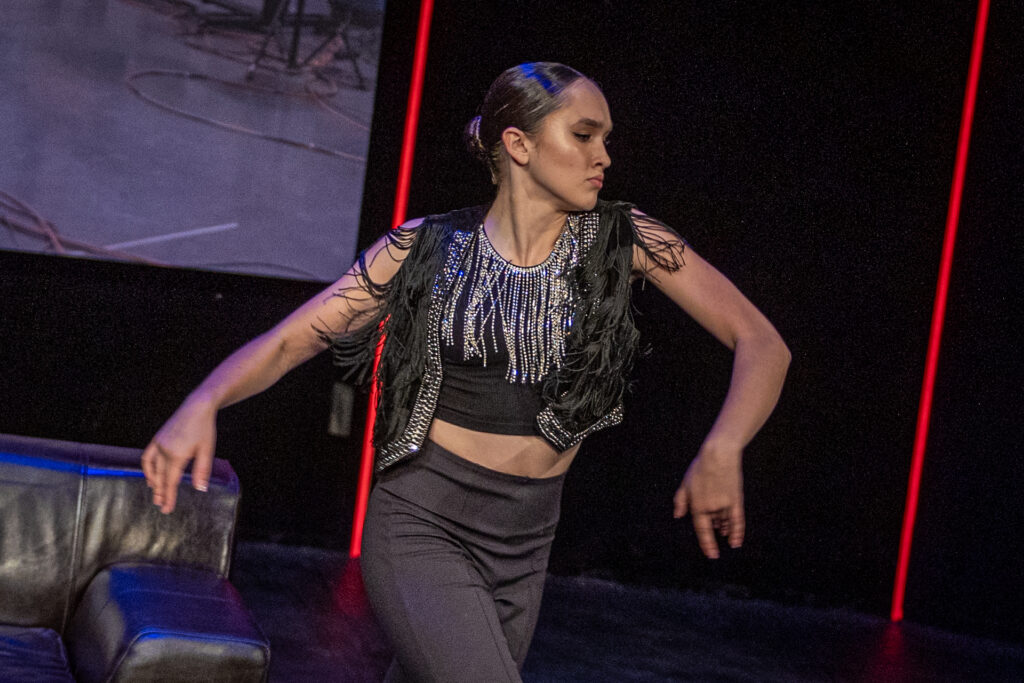 Zdjęcie. Kobieta tańczy na scenie. To brunetka z gładko zaczesanymi w kok włosami. Na sobie ma spodnie i krótką czarną bluzkę odsłaniającą brzuch. i czarną krótką kamizelkę wykończoną długimi frędzlami. Dekolt bluzki ozdabiają długie kryształowe, srebrne frędzle.