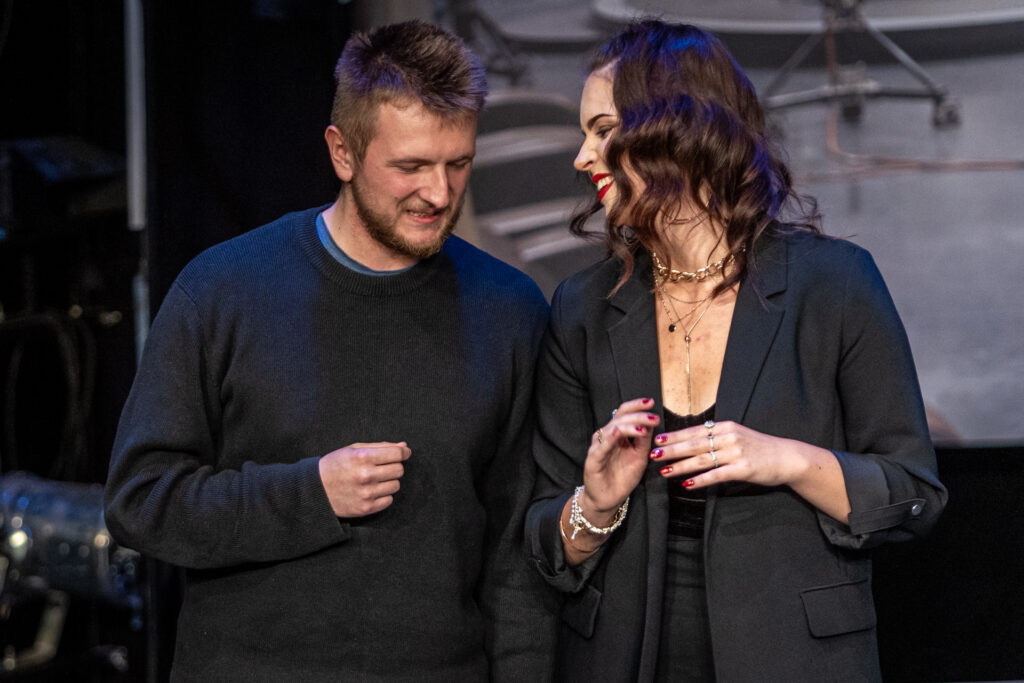 Zdjęcie. Para na scenie. Stoją obok siebie, kobieta patrzy w kierunku mężczyzny i serdecznie się uśmiecha.