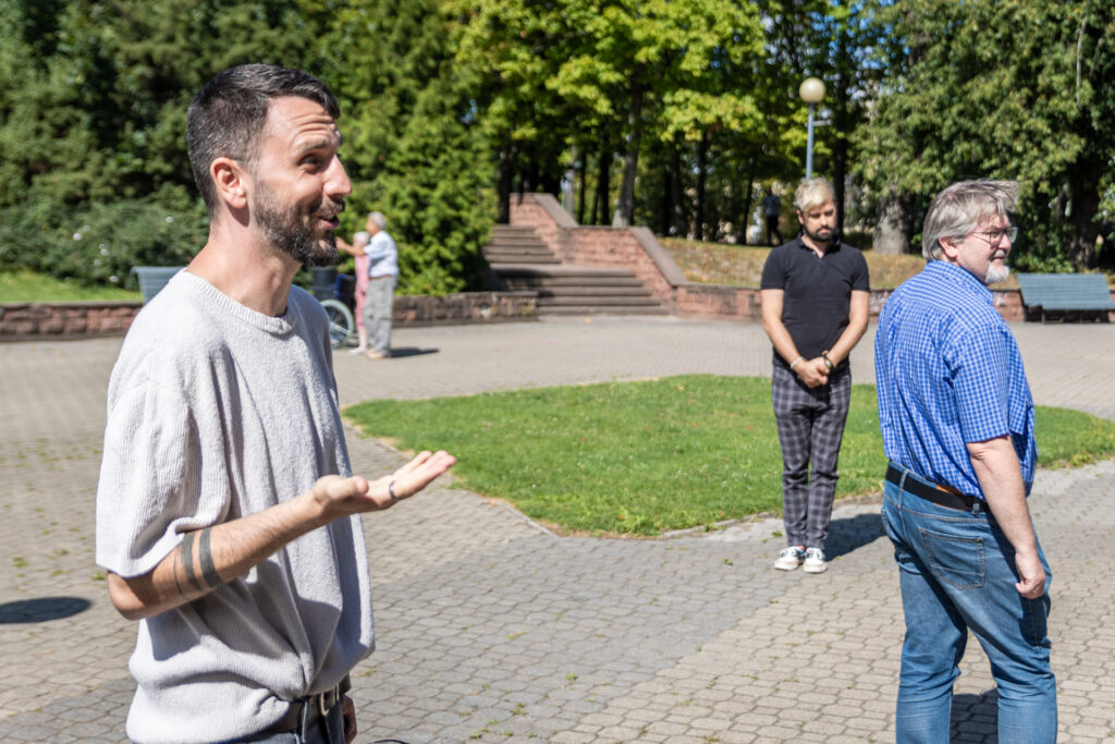 Zdjęcie. Uśmiechnięty mężczyzna z brodą tłumaczy coś. Przed nim ustawieni w różną stronę mężczyżni.