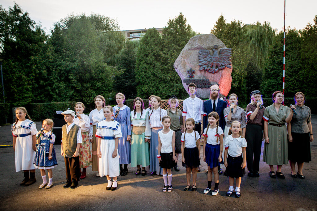 Zdjęcie. Grupa starszych i mniejszych dzieci i młodzieży stoi w kilku rzędach przed pomnikiem pamięci harcerzy.