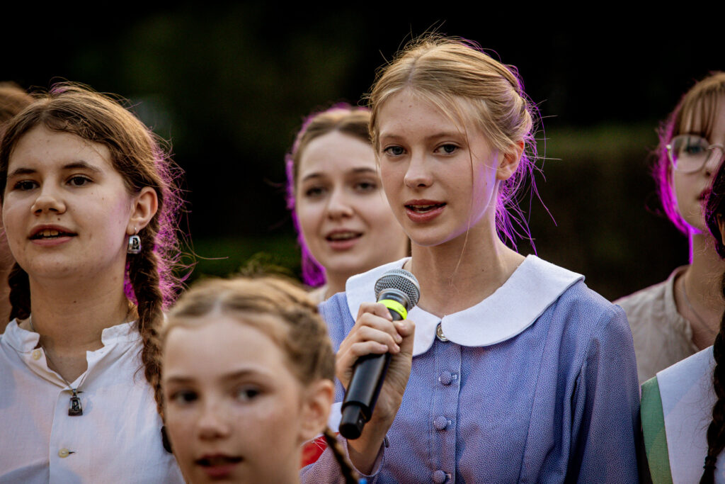 Zdjęcie. Blondynka stoi w grupie dziewczynek. Mówi do mikrofonu.