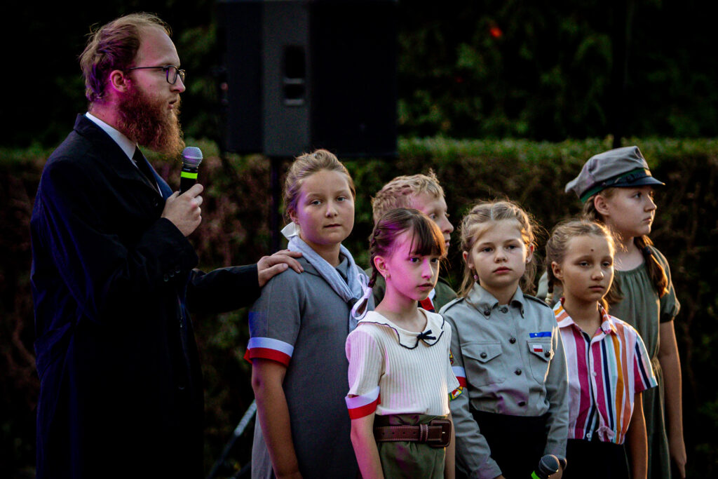 Zdjęcie. Mężczyzna o długiej brodzie trzyma za ramię dziewczynkę. Ta stoi w grupce dzieci.