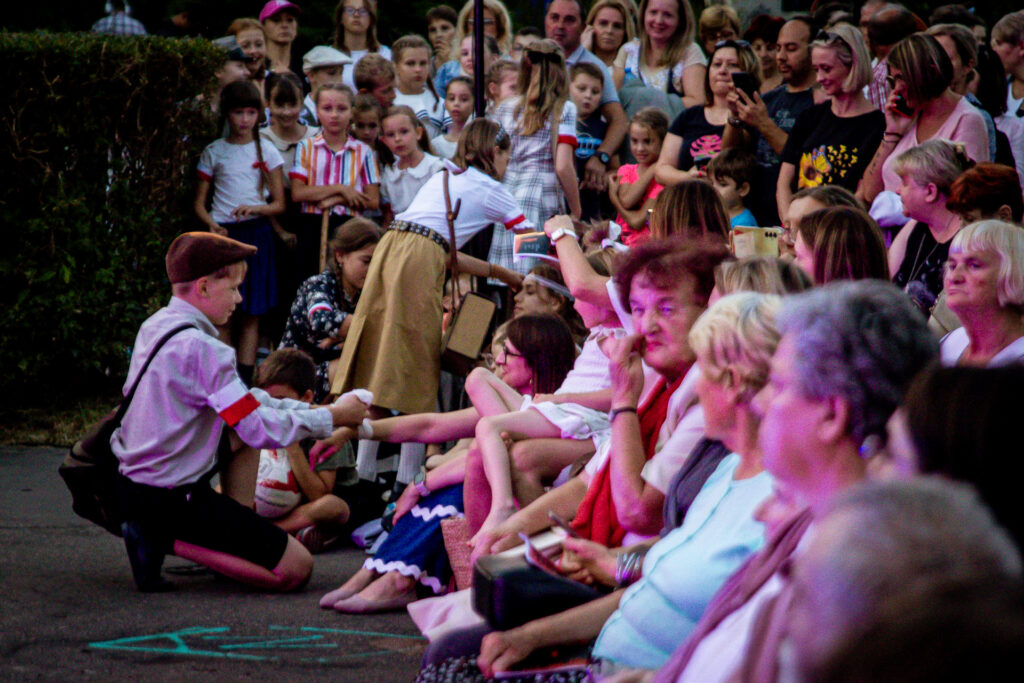 Zdjęcie. Widownia musicalu plenerowego "Kolumbowie wszechczasów". Do widzów podchodzą dzieci z biało-czerwonymi opaskami na ramionach.