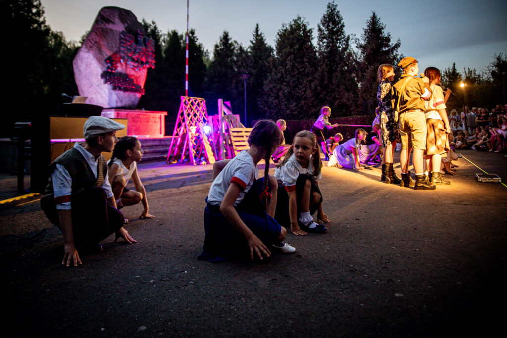 Zdjęcie. Dzieci podczas musicalu przed pomnikiem pamięci harcerzy. Grupa dzieci w strojach harcerskich śpiewa do mikrofonu. Pozostałe dzieci kucają.