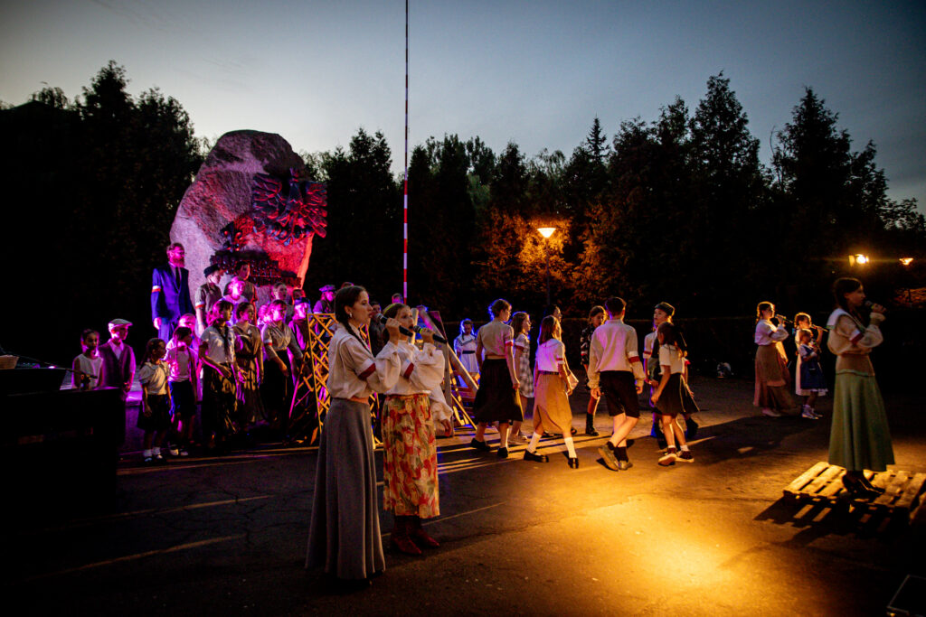 Zdjęcie. Ekipa musicalu podczas spektaklu. Przestrzeń oświetlona reflektorami, zapada zmrok.