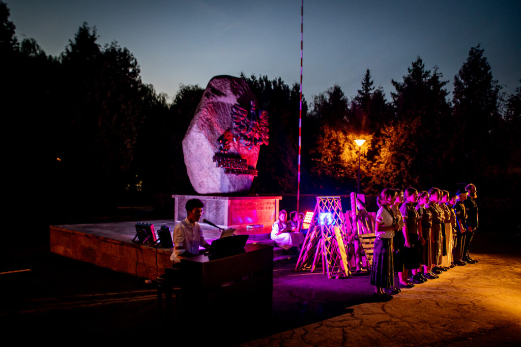 Zdjęcie. Scena musicalu plenerowego. Grupa młodzieży stoi w rzędzie. W tle pomnik-obelisk.