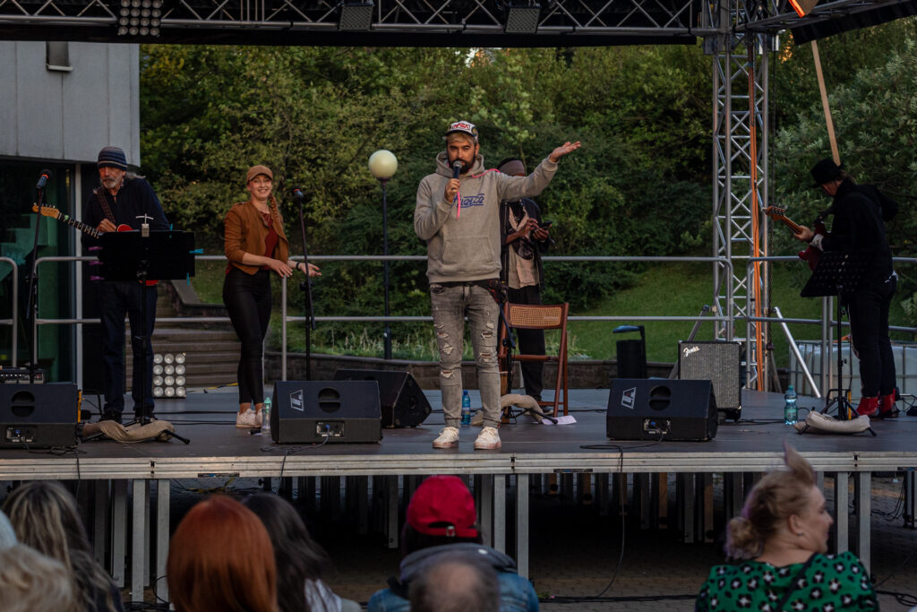 Zdjęcie. Mężczyzna w szarej bluzie z napisem Kielce mówi do mikrofonu na Letniej Scenie KCK. To wicedyrektor KCK Artur Wijata.