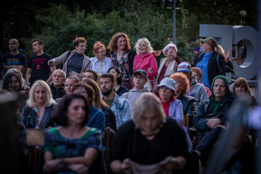 Zdjęcie. Publiczność koncertu Dobroband. Ludzie siedzą na krzesełkach. W ostatnim rzędzie stoją uśmiechnięte panie.