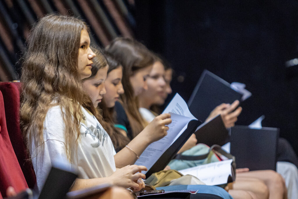 Zdjęcie. Dziewczynki siedzą w rzędzie na widowni Małej Sceny KCK. Czytają scenariusz musicalu.