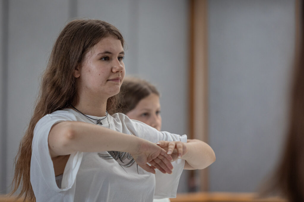 Zdjęcie. Długowłosa szatynka nastolatka patrzy przed siebie. Obie dłonie trzyma blisko siebie na wysokości piersi. Łokcie wyciąga na boki.