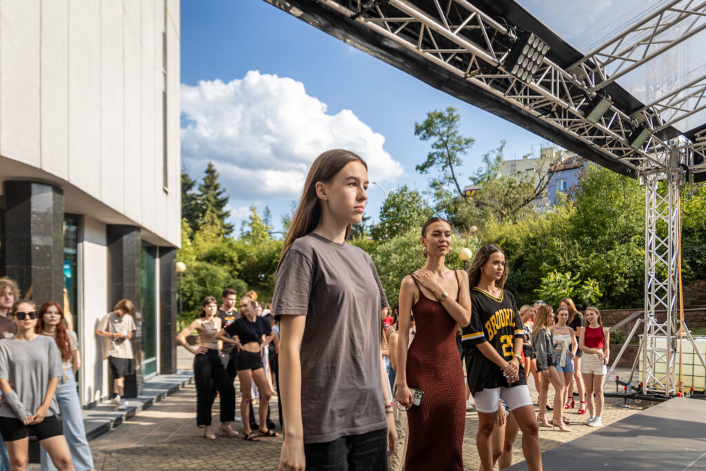 Zdjęcie. Trzy młode dziewczyny stoją na brzegu sceny letniej. W tle grupa modeli i modelek.