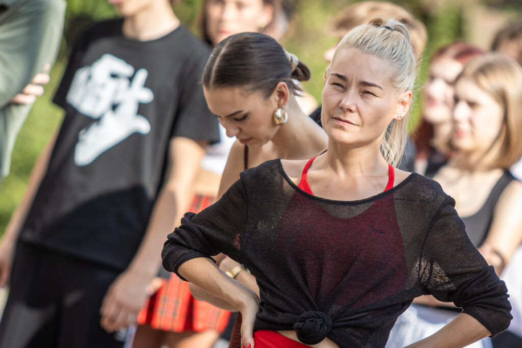 Zdjęcie. Szczupła blondynka trzyma się pod boki patrząc przed siebie. Za nią kolejne modelki.