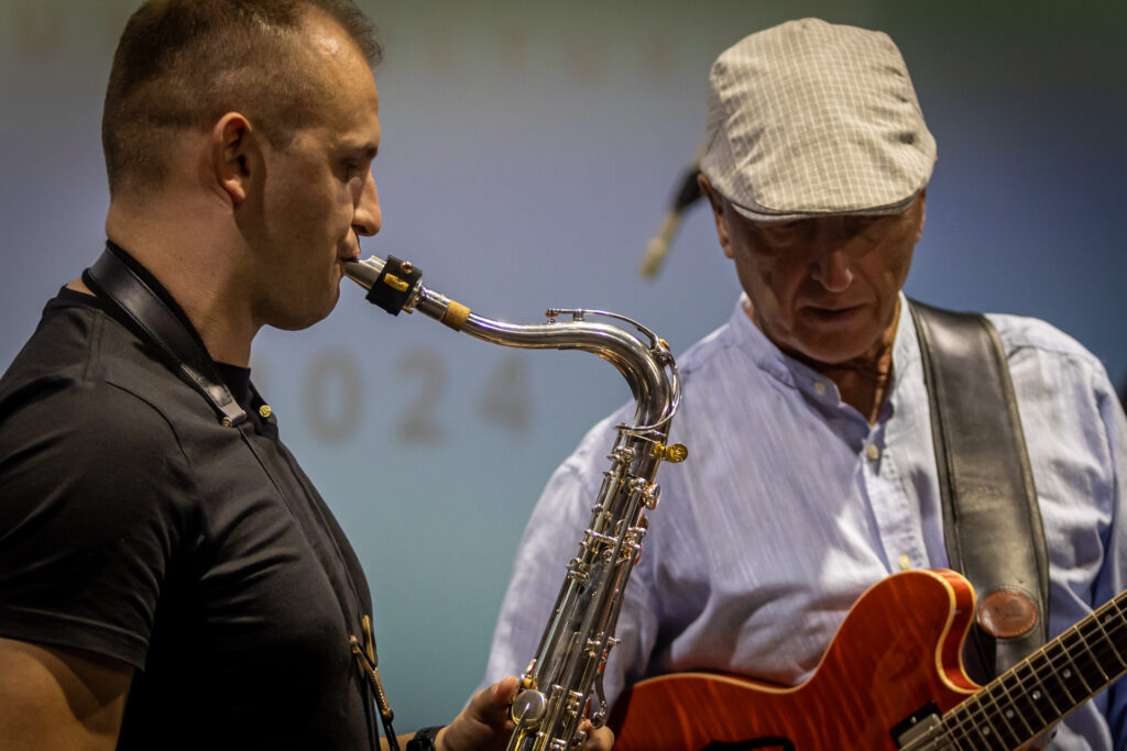 Zdjęcie. Dwaj mężczyźni grają na instrumentach. Jeden na gitarze, drugi na saksofonie.