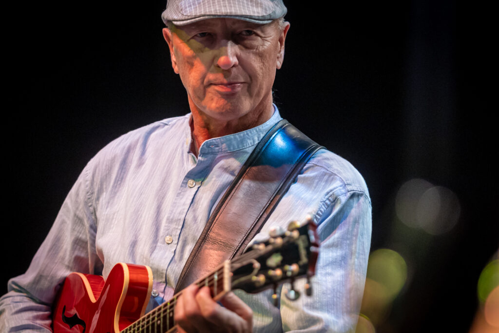 Zdjęcie. Starszy mężczyzna gra na gitarze. Na głowie ma czapkę z daszkiem typu kaszkiet.