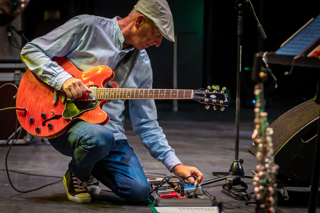 Zdjęcie. Mężczyzna z gitarą klęczy na jedno kolano. Przekręca dłonią pokrętła miksera, leżącego na podłodze.